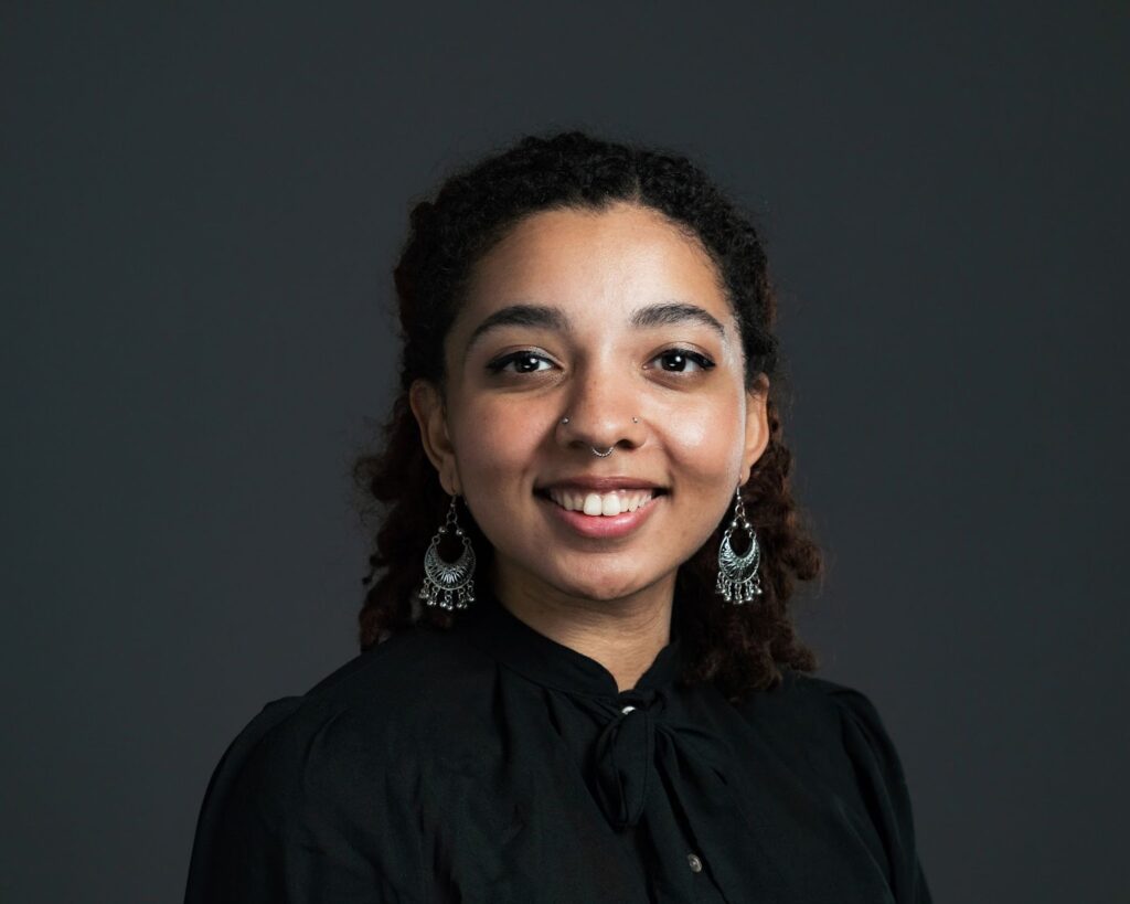 A smiling woman with medium brown skin, long black hair, and a black shirt. She has a nose ring and piercing and is wearing large dangly earrings.