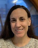 A smiling woman with fair skin and long brown hair. She is wearing a white sweater with black specks.