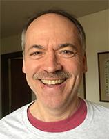 A balding fair-skinned man with a brown mustache and a broad smile. He is wearing white and red t-shirts.