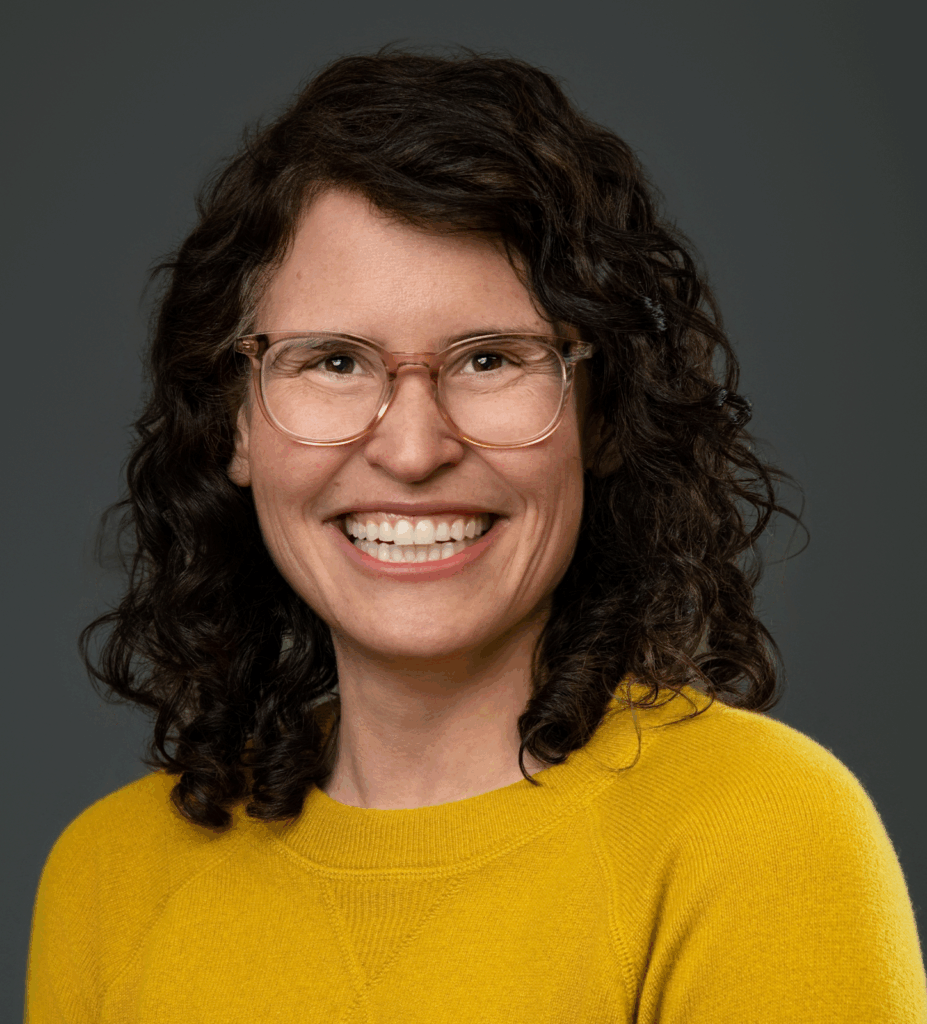A woman with light skin, brown eyes and curly dark hair wearing a yellow sweater and glasses grins broadly.