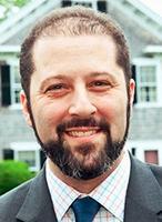 A fair-skinned man with dark hair, mustache, and beard smiles at the camera; he is wearing a checked shirt, blue tie, and gray blazer.