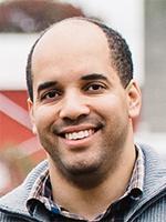 A man of medium dark complexion with short cropped hair smiles; he is wearing a checked shirt and grey cardigan