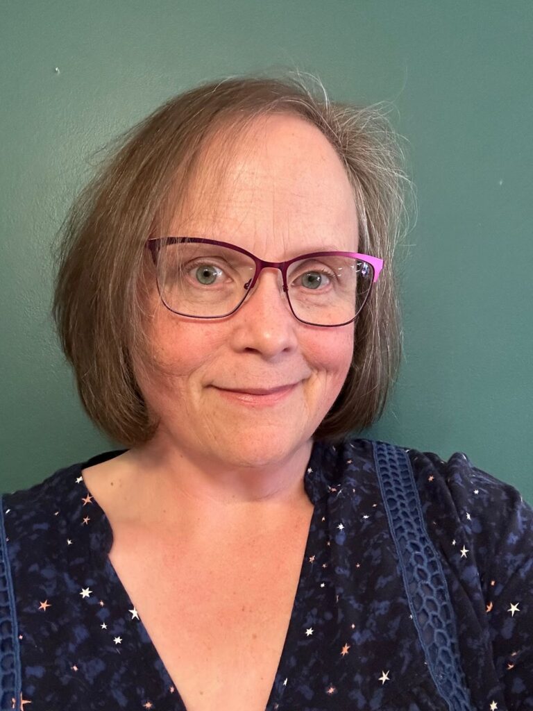 A woman with pinkish complexion, short auburn hair, wearing glasses and a blue shirt with stars on it.