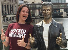 A smiling woman with medium light complexion and long brown hair giving two thumbs up next to a statue of the Fonz, who also has two thumbs up. The woman is wearing a maroon shirt that reads WICKED SMAAHT in white letters.